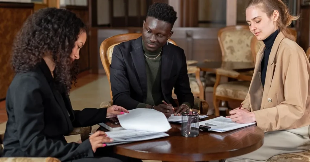 Three professionals reviewing documents during customer acquisition due diligence meeting at conference table.