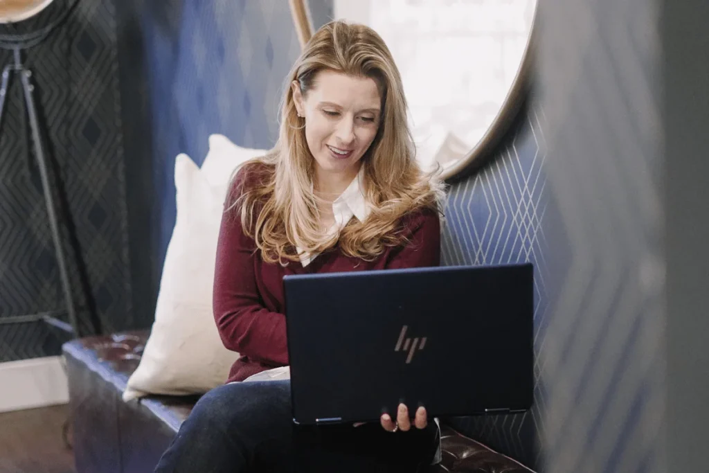 Nora Sudduth seated on a leather bench works on an HP laptop, smiling.