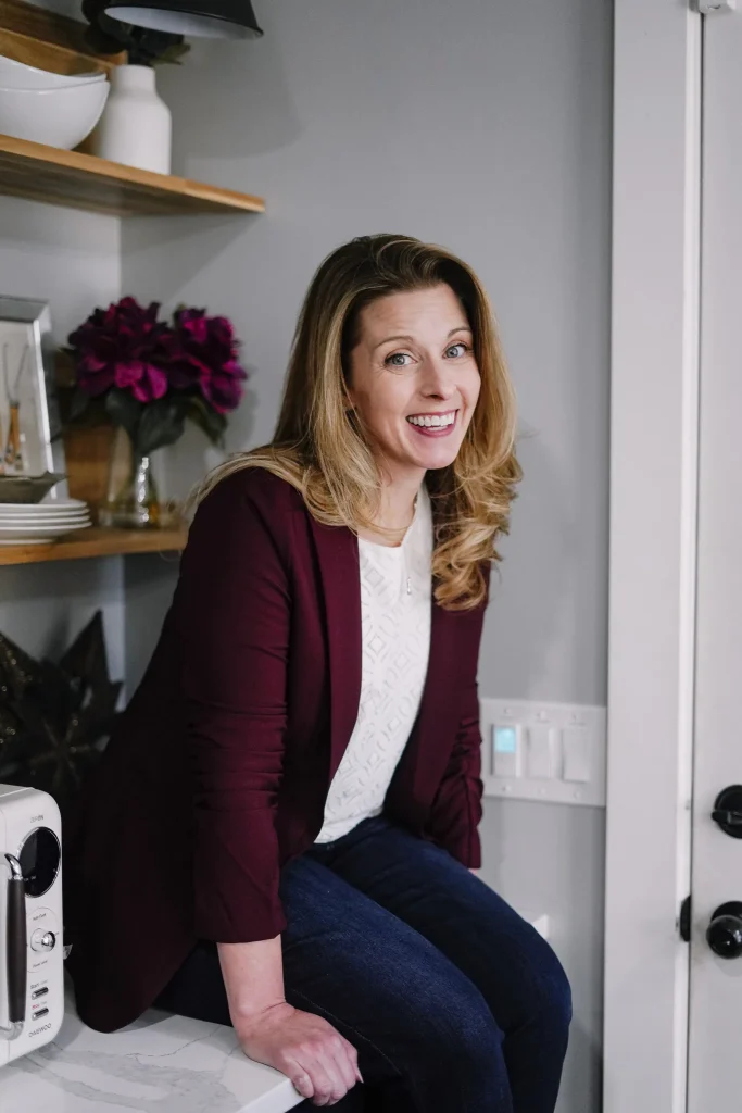 Nora Sudduth in a burgundy blazer and jeans sits on a kitchen counter, with shelves holding dishes and flowers in the background, creating a warm, cheerful atmosphere.